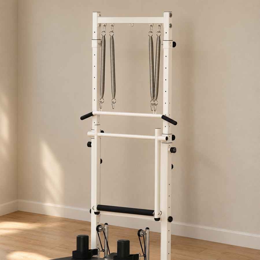 A white and black multi-functional pilates tower rack with pull-up bars and hanging resistance bands, set against a beige wall and light wood floor.