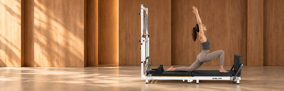 A woman in a gray workout outfit performs a Pilates pose on the OUBLANC RT-101 Pilates reformer machine inside a modern studio with warm wood-paneled walls and floor.