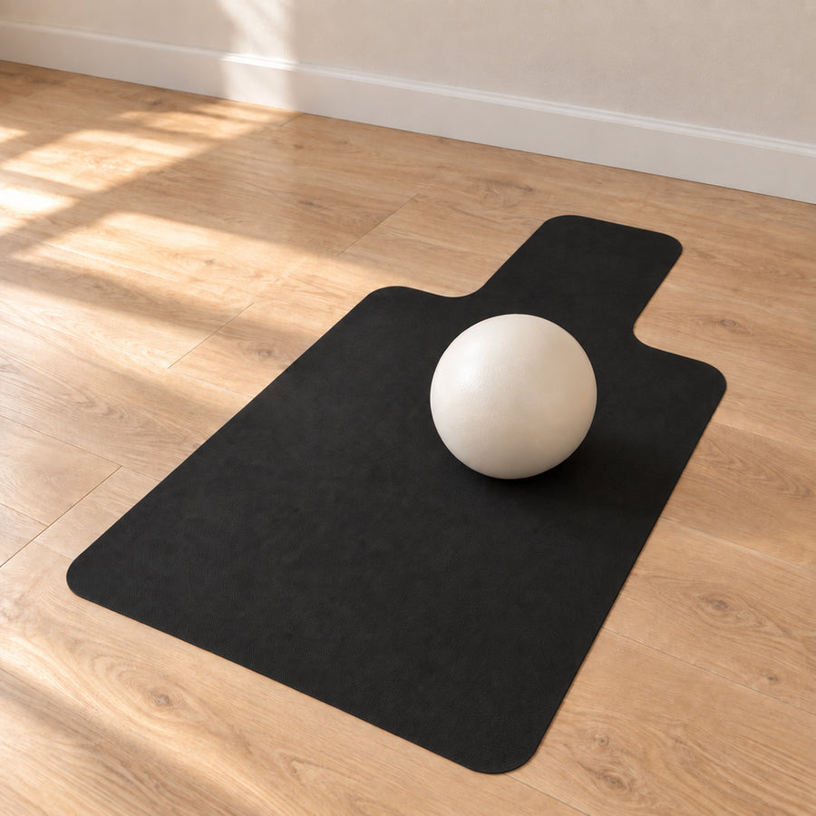 A black reformer mat and a Pilates ball, displayed on a light wood floor with a white sphere prop, in a room setting with warm sunlight and shadows.