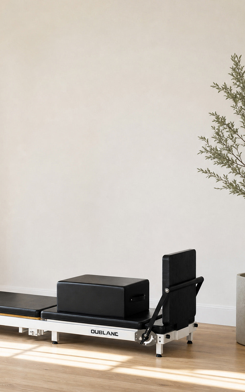 A black and white Pilates reformer machine with a black sitting box on a wooden floor against a beige wall, with a potted plant on the right.