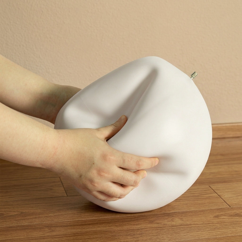 A pump nozzle is inserted into a Pilates ball to deflate it. Meanwhile, a person's hands are pressing the ball to speed up deflation on a wooden floor against a beige wall.