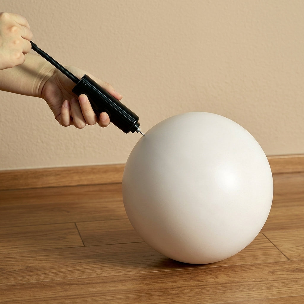 A hand using a black handheld pump to inflate a light gray Pilates ball on a wooden floor against a beige wall.