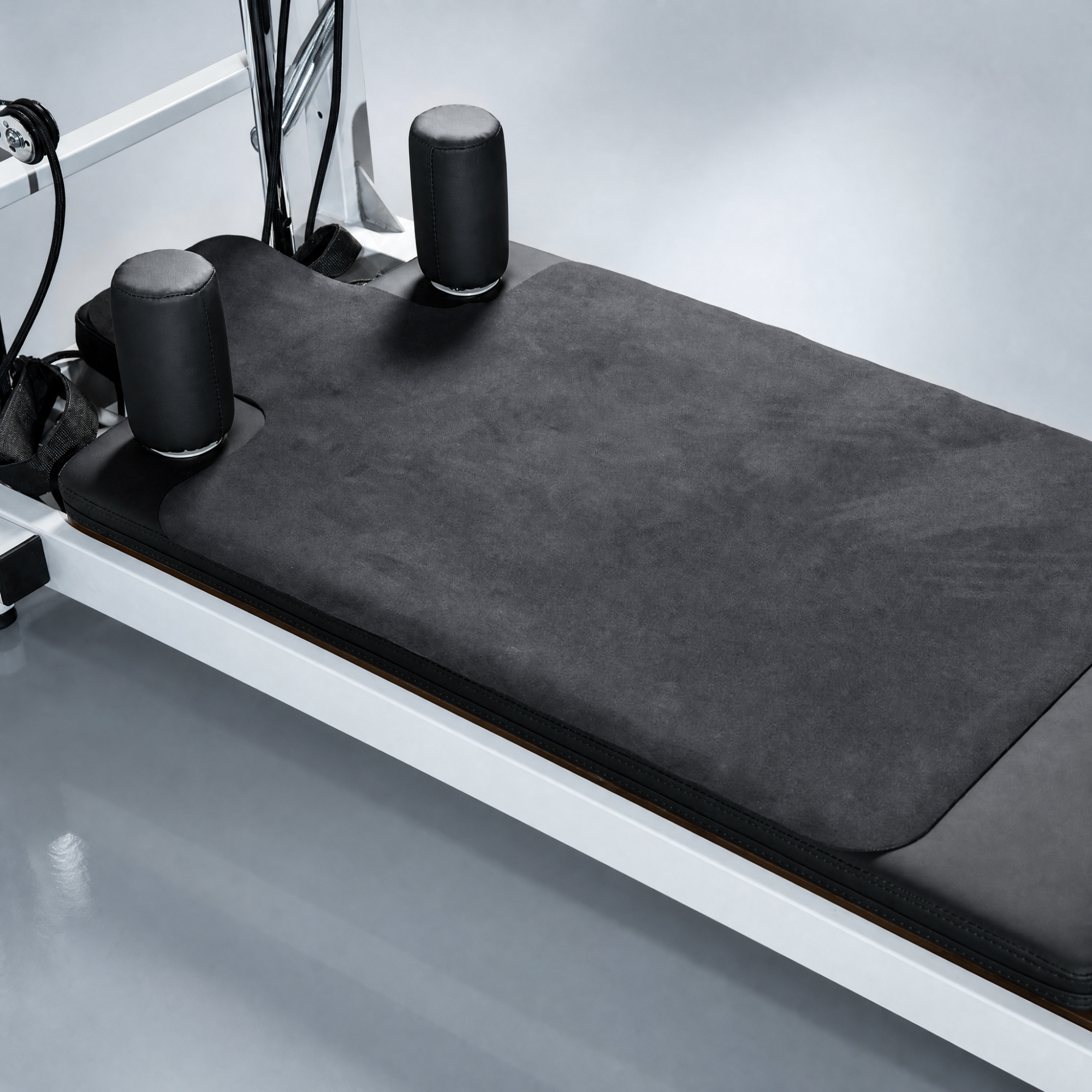 Pilates reformer machine with a black textured mat, black padded shoulder rests, and a white metal frame on a light gray studio floor.