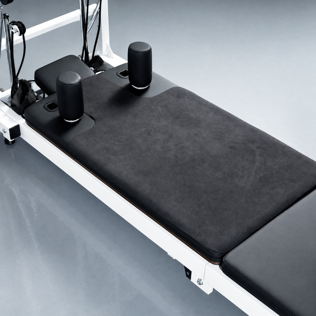 Pilates reformer machine with a black textured mat, black padded shoulder rests, and a white metal frame on a light gray studio floor.