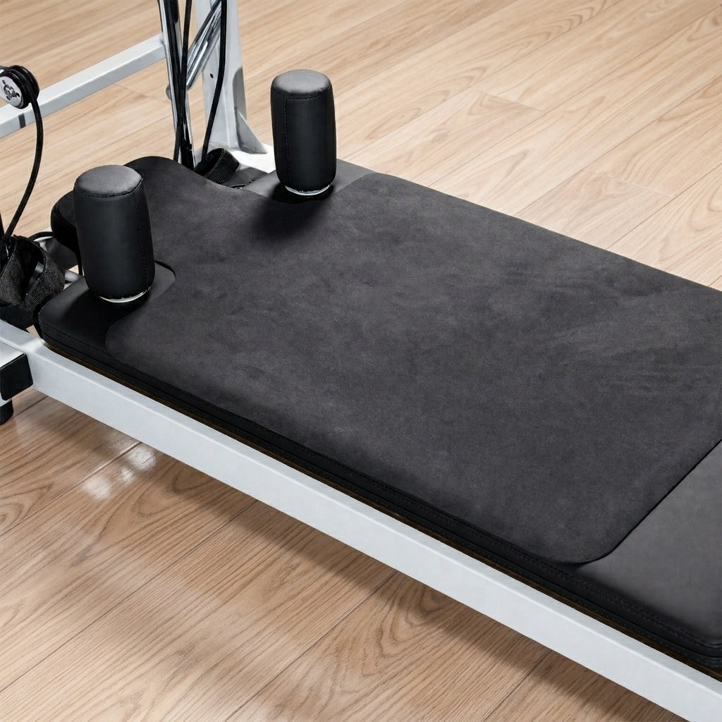 Pilates reformer machine with a black reformer mat on the carriage, set on a light wood floor.