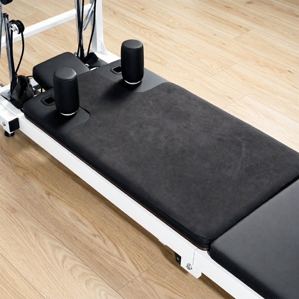 Pilates reformer machine with a black reformer mat on the carriage, set on a light wood floor.