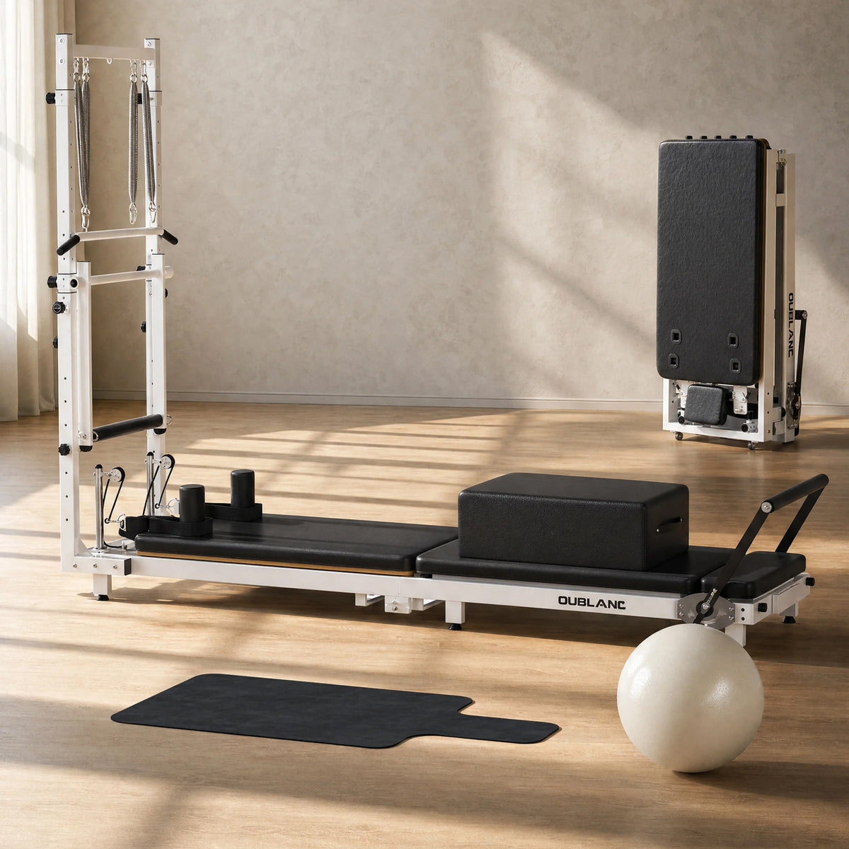 OUBLANC RT-101 Pilates reformer machine with a white frame and black padding, shown in a bright room with wood floors. A black reformer mat, a light gray Pilates ball, and a folded version of the RT-101 are arranged nearby.