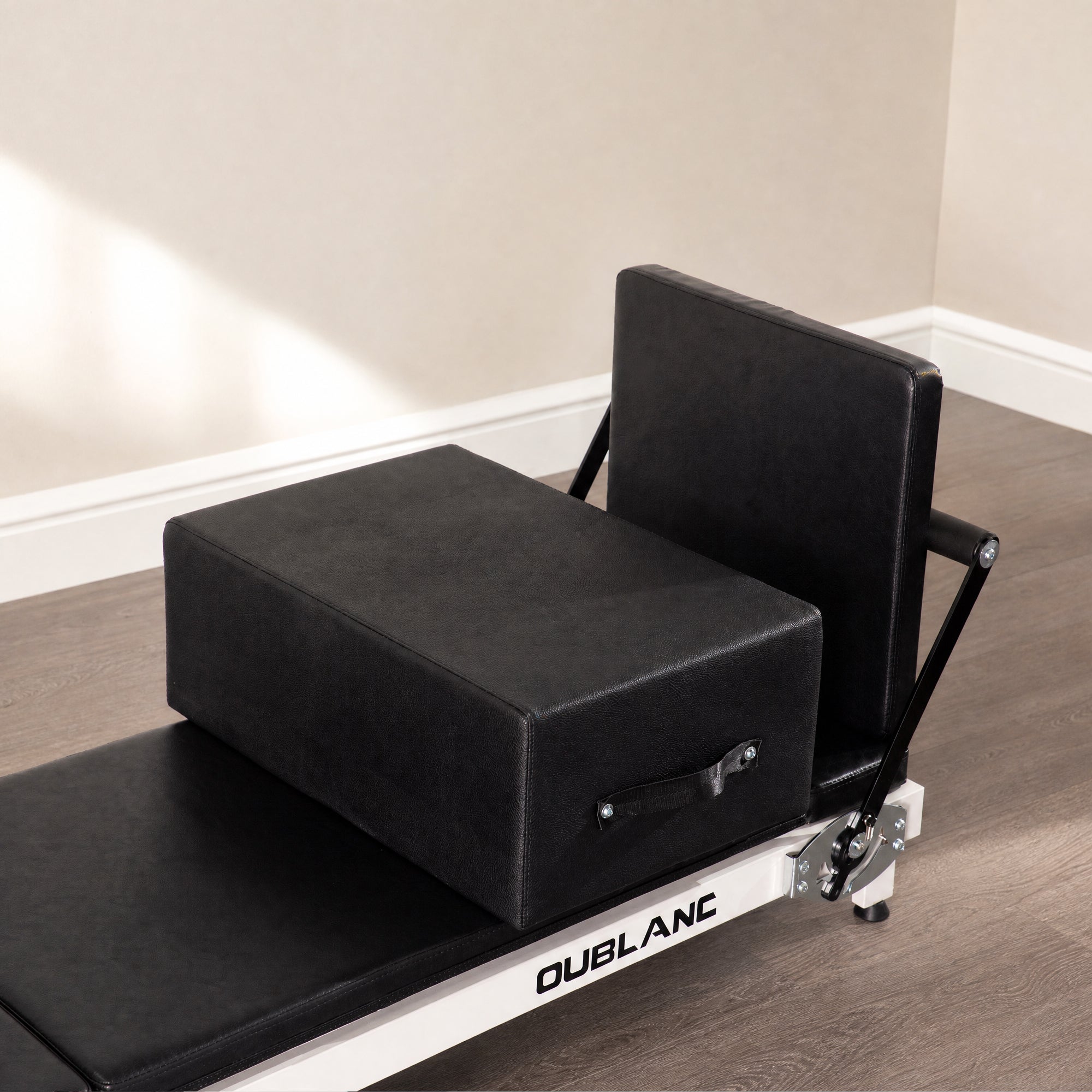 A black Pilates reformer machine with a rectangular sitting box on top, shown in a studio setting with wood flooring and beige walls. The brand name 'OUBLANC' is visible on the frame.